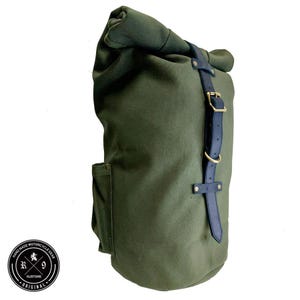 Op de afbeelding: Olijfgroene rugzak met oprolbare bovenkant, zwarte leren riem en gouden gesp. De tas heeft een zijvak en een rond logo met "HAND MADE MOTORCYCLE GEAR" en "KUSTOMS ORIGINAL".