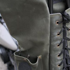 Op de afbeelding: Close-up van een groene canvas tas met een zak en zwarte veters. De tas heeft een versleten, getextureerde uitstraling met metalen ogen en een ronde patch. De tas is bevestigd aan een motorfiets.