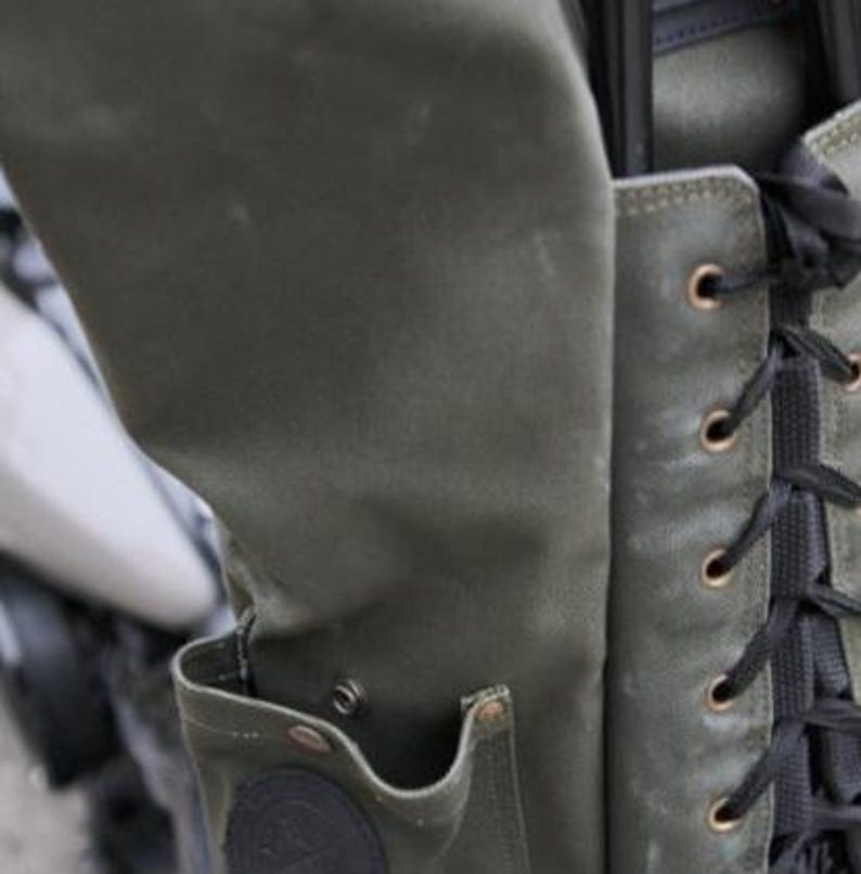 Op de afbeelding: Close-up van een groene canvas tas met een zak en zwarte veters. De tas heeft een versleten, getextureerde uitstraling met metalen ogen en een ronde patch. De tas is bevestigd aan een motorfiets.