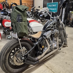 Op de afbeelding: Een zwarte custom motorfiets met een vlammenontwerp op de brandstoftank en een bruin leren zadel. Hij heeft een grote achterband, een groene canvas tas en een zwarte waterfles. De uitlaatpijpen zijn gewikkeld in een tan materiaal.