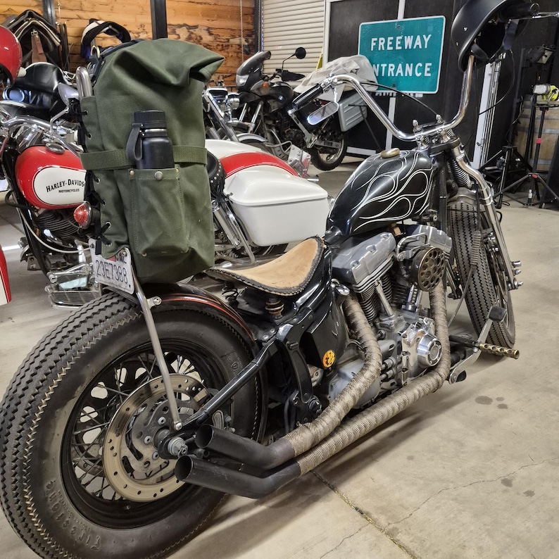 Op de afbeelding: Een zwarte custom motorfiets met een vlammenontwerp op de brandstoftank en een bruin leren zadel. Hij heeft een grote achterband, een groene canvas tas en een zwarte waterfles. De uitlaatpijpen zijn gewikkeld in een tan materiaal.