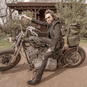 Op de afbeelding: Een vintage zwarte motorfiets met een vlammenontwerp op de tank. De motorfiets heeft een grote koplamp, hoge stuurstangen en een groene canvas tas aan de achterkant. De bestuurder draagt een zwarte jas en laarzen.