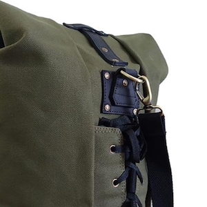 Op de afbeelding: Olijfgroene canvas tas met zwarte leren details en een zwarte schouderriem. Het heeft een decoratieve leren patch met messing hardware en een zijvak met zwarte veters.