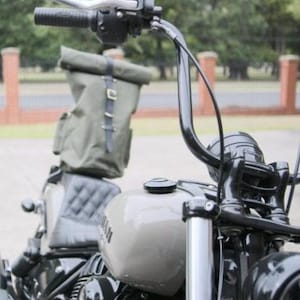 Op de afbeelding: Close-up van een motorfiets met een grijze brandstoftank en zwarte stuur. Een groene canvas rugzak is met zwarte riemen aan de achterbank bevestigd. De motorfiets heeft een zwarte koplamp en een gewatteerde zwarte zitting.