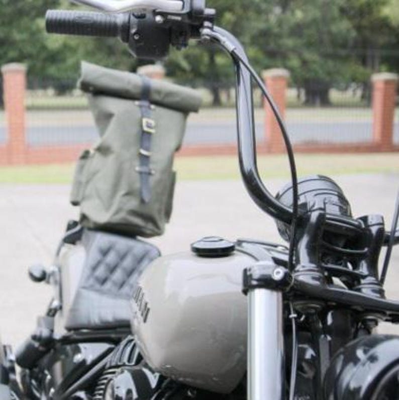 Op de afbeelding: Close-up van een motorfiets met een grijze brandstoftank en zwarte stuur. Een groene canvas rugzak is met zwarte riemen aan de achterbank bevestigd. De motorfiets heeft een zwarte koplamp en een gewatteerde zwarte zitting.