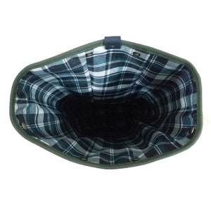 Op de afbeelding: Een open, olijfgroene tas met een donker geruit interieur. De tas heeft een donker leren riem en metalen ogen rond de bovenkant. Het ruitpatroon heeft tinten blauw, groen en wit.