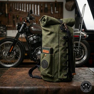 Op de afbeelding: Een groene canvas roll-top rugzak met zwarte accenten, leren details en een voorvak. De rugzak heeft een "HAND MADE" patch en een rond logo. Een motorfiets is op de achtergrond te zien.