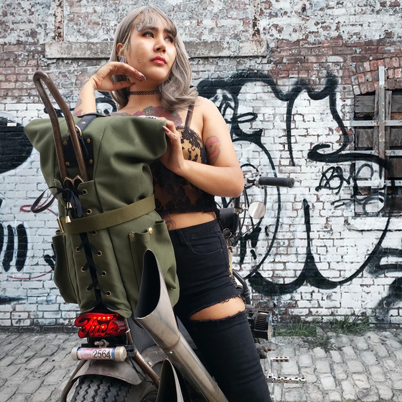 Op de afbeelding: Een olijfgroene motorzadeltas met een veter aan de voorkant en meerdere zakken. De tas is gemonteerd op een aangepaste motorfiets met een rood achterlicht. De achtergrond is een bakstenen muur met graffiti.