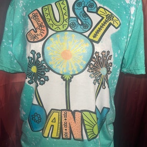 May include: A turquoise blue t-shirt with a white bleach design. The design features the words "JUST DANDY" in a colorful, whimsical font with dandelion illustrations.