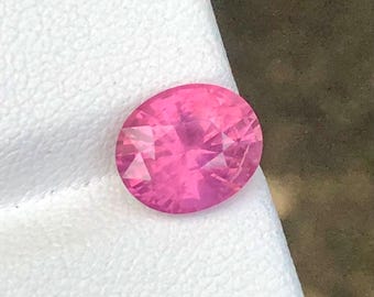 May include: A faceted, oval-cut pink gemstone. The gem is a vibrant shade of pink, with multiple facets that reflect light. The gemstone is set against a white surface, creating a contrast that highlights its colour and brilliance.