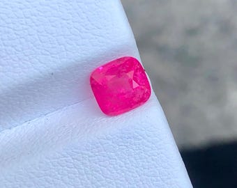 Puede incluir: Una gema rosa intenso, talla cojín. La gema está colocada sobre una superficie blanca y texturizada. Las facetas de la gema reflejan la luz, mostrando su brillo. Esta imagen resalta la belleza y el color de la piedra preciosa.