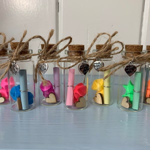 May include: Eight small glass jars with cork tops, each containing a folded origami shape and a rolled up note. Each jar has a silver charm with a heart and the text "Made with love". The jars are arranged in a row on a light blue surface.