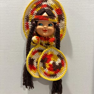 May include: A vintage doll with a ceramic face and crocheted clothing. The doll has brown braids, a yellow, red, and brown headdress, and matching circular accents. The doll's face has blue eyes and a red mouth. The doll is a wall hanging.