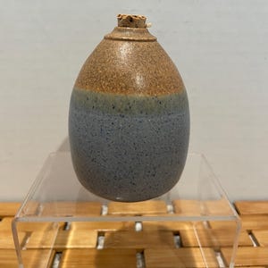 May include: A handmade ceramic bottle with a cork stopper. The bottle has a rounded shape and is divided into three sections: a brown top, a blue-grey middle, and a thin green-blue band between them. The bottle is displayed on a clear acrylic stand.