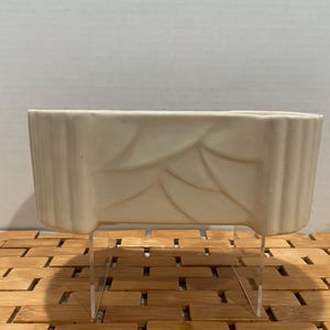 May include: White ceramic planter with a leaf design and clear acrylic legs. The planter is sitting on a bamboo mat.