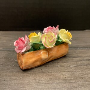May include: A small, wooden planter with a log-like design, filled with five porcelain roses in shades of pink and yellow.