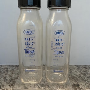 May include: Two clear glass baby bottles with black caps and blue labels that read "DAVOL "ANTI-COLIC" BRAND Nurser PAT. PEND."