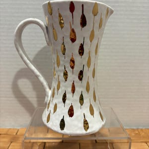 May include: White ceramic pitcher with a textured surface and a curved handle. The pitcher is decorated with gold, brown, and orange leaf-shaped designs. It is displayed on a clear acrylic stand.