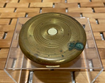 May include: A vintage, round brass compact with a concentric circle design. The compact has a small, off-white center and some green discoloration. It sits on a clear acrylic stand.