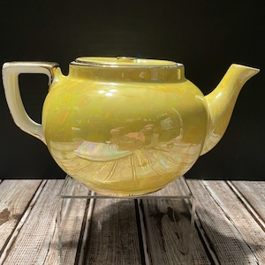 May include: A yellow teapot with a pearlescent glaze. The teapot has a round body and a spout on the right side. The handle is on the left side and is white with a gold trim.