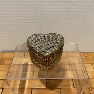 May include: A heart-shaped, vintage-style trinket box. The box is made of gold-colored metal with a perforated lid and sides. The box is sitting on a clear acrylic stand.