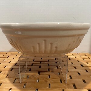 May include: A cream-colored ceramic mixing bowl with a wide rim and vertical grooved detailing around the exterior. The bowl is displayed on a clear acrylic stand, set against a neutral background.