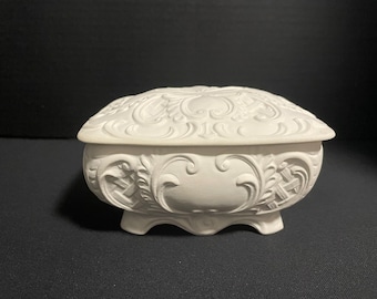 May include: A white trinket box, oval in shape, with a lid. The box is decorated with elaborate, raised floral and scrollwork patterns. It stands on three small feet and is made of a solid, opaque material.