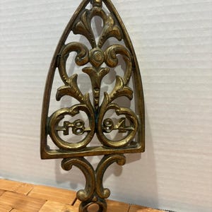 May include: Ornate, antique brass trivet with a decorative openwork design. The trivet is shaped like an iron and features the numbers "18" and "94". The handle has a circular opening. The trivet is a warm, aged gold color.