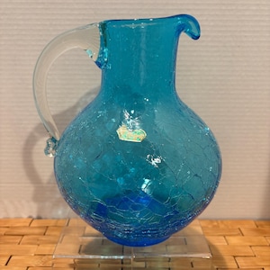May include: A turquoise crackle glass pitcher with a clear glass handle. The pitcher has a rounded body and a spout. A small sticker is affixed to the front. The pitcher is displayed on a clear acrylic stand.