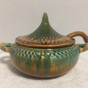 May include: A green and brown ceramic serving dish with a lid. The lid has a pointed top and a handle. The dish has two handles on the sides.