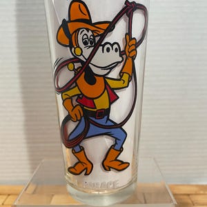May include: Clear glass with a cartoon cowboy character illustration. The cowboy is wearing an orange hat, vest, and boots, a yellow shirt, and blue pants. The cowboy is holding a red lasso. The word "GARAGE" is printed at the bottom.