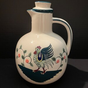 May include: White ceramic pitcher with a green lid and a hand-painted rooster design. The rooster is green, blue, yellow, and red. The pitcher has a green and white floral design.