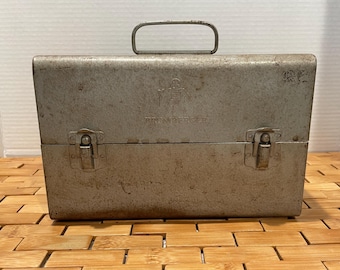 May include: A vintage metal toolbox with a handle and two latches. The toolbox is a weathered silver color with the word "PRUMBERGER" printed on the front. The toolbox is sitting on a wooden surface.