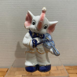 May include: A white ceramic elephant figurine playing a blue violin. The elephant wears a blue bow tie, a white suit with gold trim, and blue shoes. The elephant's ears and trunk are pink.
