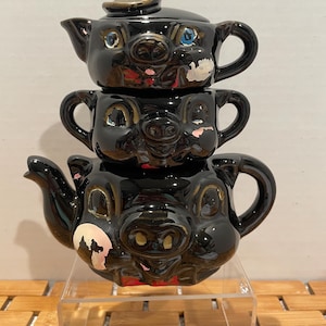 May include: A set of three black ceramic teapots shaped like pig faces. The teapots are stacked on top of each other, with the smallest teapot on top. The teapot on top has a brown hat. Each teapot has painted details, including eyes, a nose, and a mouth.