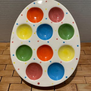 May include: White ceramic egg-shaped serving dish with nine colorful circular indentations. The indentations are filled with red, pink, yellow, blue, and green. Small dots of various colors are scattered around the dish.