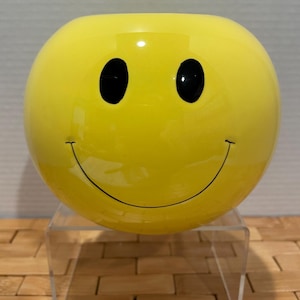 May include: A round, yellow smiley face vase with black eyes and a curved black mouth. The vase is glossy and sits on a clear acrylic stand. The smiley face design is a classic and cheerful decorative item.