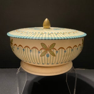 May include: A vintage white and blue striped tin container with a floral design and a gold knob on the lid. The container is on a clear acrylic stand.