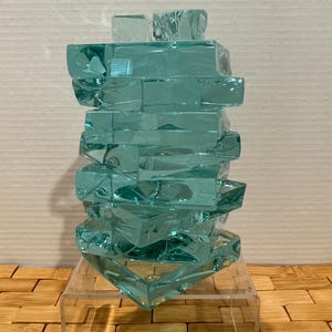 May include: A teal-colored glass sculpture composed of stacked rectangular blocks, creating a textured, abstract form. The sculpture sits on a clear acrylic stand, showcasing its geometric design. The piece is displayed on a wooden surface.