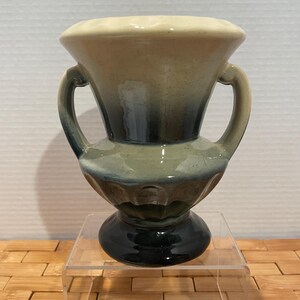 May include: A small, decorative ceramic vase with two handles. The vase features a cream-colored rim, transitioning to shades of blue and green, and a dark base. It is displayed on a clear acrylic stand.