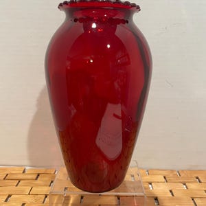 May include: A ruby red glass vase with a flared rim and a rounded body. The vase has a smooth, glossy finish and sits on a clear acrylic stand. The rim has a decorative, scalloped edge.