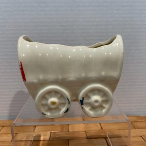May include: A cream-colored ceramic wagon planter with two wheels on each side. The wagon has a rectangular shape with a curved top and a red design on the side. The wheels have yellow and blue accents.