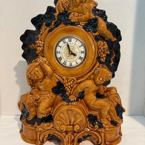 May include: A ceramic mantel clock featuring a tan finish with dark brown accents. The clock face has black Roman numerals and a gold center. The frame is decorated with three sculpted figures of cherubs and clusters of grapes and leaves.