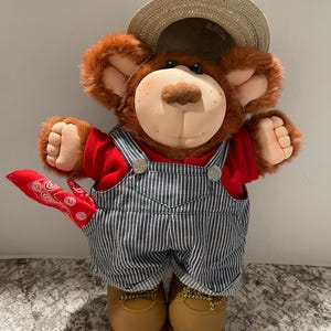 May include: A plush bear doll wearing a red shirt, blue and white striped overalls, and brown work boots. The bear has a straw hat and a red bandana tied around its neck. The bear's fur is brown and its face is tan.