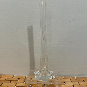 May include: A tall, clear glass vase with a flared base and a slender, elongated neck. The vase is displayed on a clear acrylic stand, showcasing its elegant design. The vase's shape is reminiscent of a flower stem.