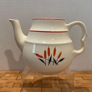 May include: Cream-colored ceramic teapot with a rounded body, a curved spout, and a unique handle. The teapot features a red rim and a red stripe around the middle. It is decorated with orange cattails and black leaves.