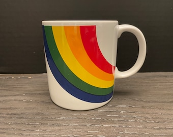 May include: White ceramic mug with a rainbow design. The rainbow is a half circle with red, orange, yellow, green, blue, and indigo stripes.