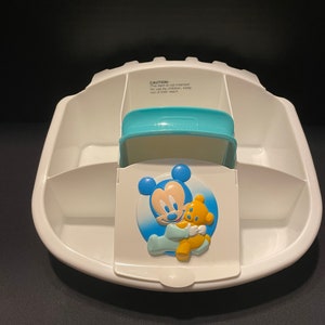 May include: White plastic baby food storage container with four compartments. The container has a blue lid with a Mickey Mouse and a teddy bear design. The lid has a safety warning that reads "CAUTION: This item is not intended for use by children, keep out of their reach."