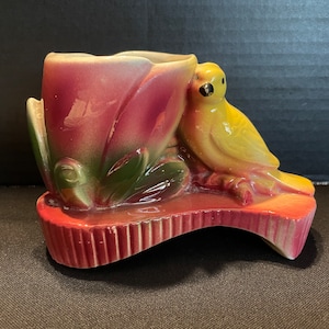 May include: A vintage ceramic planter shaped like a flower with a yellow bird perched on the side. The planter is decorated with pink, green, and brown glaze.