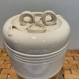 May include: White ceramic jar with a lid featuring a decorative swirl design. The jar has a cylindrical shape and is decorated with horizontal lines.